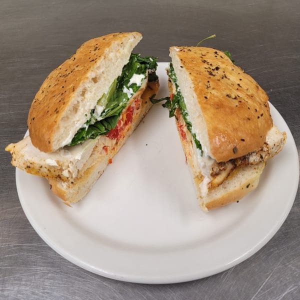 A grilled chicken sandwich on focaccia bread, cut in half, filled with greens, tomatoes, and cheese.