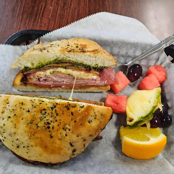 A pesto focaccia sandwich with melted cheese and deli meat, served with watermelon, pineapple, and orange slices.
